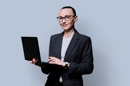 Middle-aged business serious woman using laptop on gray background. 30s successful female teacher mentor manager worker employee director looking at camera. Internet online technologies work teaching