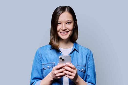 Young happy woman using smartphone in gray background. Smiling 20s female looking at camera texting. Mobile Internet apps applications technologies for work education communication shopping healthcare