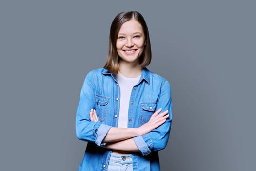 Young beautiful confident woman with crossed arms on gray studio background. Smiling attractive 20s female in denim looking at camera. Lifestyle, fashion beauty style, youth business work education