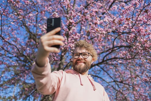 Happy curly man takes selfie against backdrop of flowering tree in spring for his internet communications. Weekend and social networks