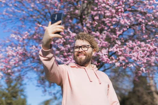 Happy curly man takes selfie against backdrop of flowering tree in spring for his internet communications. Weekend and social networks