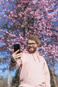 Happy curly man takes selfie against backdrop of flowering tree in spring for his internet communications. Weekend and social networks