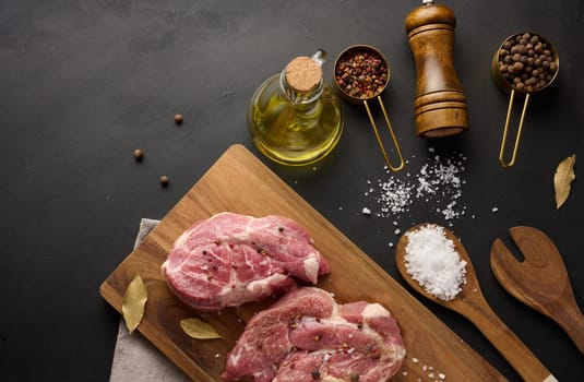 Two raw pork neck steaks on a board and spices for cooking. Top view of black table