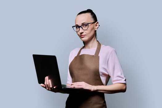 Portrait of 30s serious confident woman in an apron using laptop, looking at camera on grey studio background. Worker, startup, small business, job, service sector, staff, youth concept