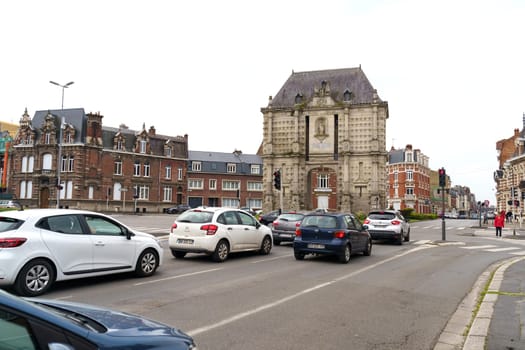 Cambrai, France - May 21, 2023: A line of cars driving down a city street bordered by towering buildings on both sides.
