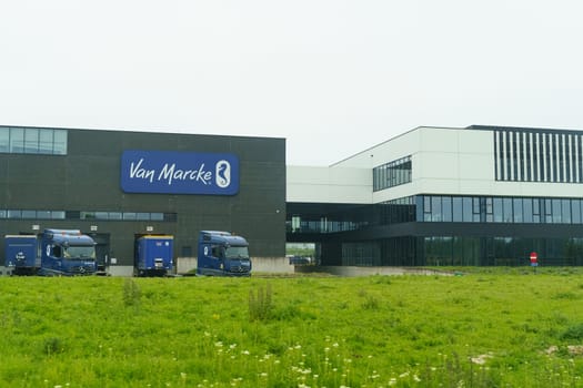 Ronchin, France - May 22, 2023: Van Marcke company trucks parked outside the brands modern warehouse on an overcast day, with a lush green field in the foreground.