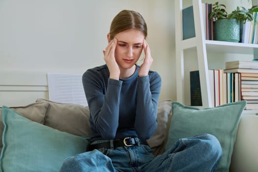 Young suffering teenage female having headache, holding head with hands, sitting on couch at home. Health, problems, mental difficulties, depression, youth concept