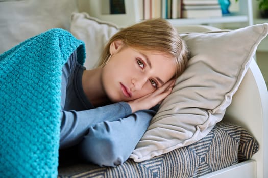 Relaxed resting young teenage female, looking at camera, lying on pillow under blanket, at home on couch. Health, beauty, rest calm peace, lifestyle, youth concept