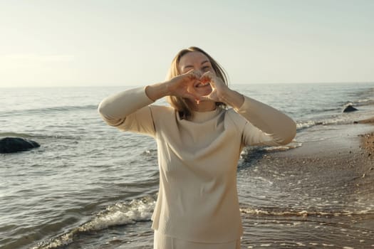 A woman stands on the shore, playfully depicting a heart with her hands, against the backdrop of gentle waves and a clear sky.