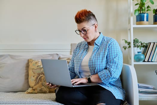 Middle aged woman sitting on couch at home using laptop. Serious female with red haircut in casual clothes working on computer, relaxing, communicating