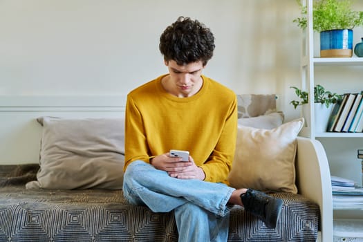 Young guy holding smartphone in hands sitting on couch at home. Handsome university college student 19-20 years old in yellow with curly hair. Technologies, mobile apps, study communication leisure