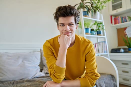 Portrait of handsome smiling guy looking at camera, web cam view, sitting on couch at home. Young man in yellow with curly hair, university college student. Lifestyle, youth 19-20 years old concept