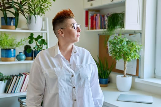 Profile portrait of serious middle-aged woman in glasses with red haircut looking at window in home interior, copy space. Mature people, lifestyle, health, life concept