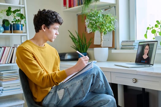 Guy college student sitting at home at desk looking at laptop screen, making notes in notebook. Young male studying online, watching webinar, browsing Internet. E-learning, education, technology youth