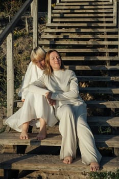 Mom and daughter are sitting on a wooden staircase in the rays of the setting sun.