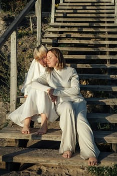 Mom and daughter are sitting on a wooden staircase in the rays of the setting sun.