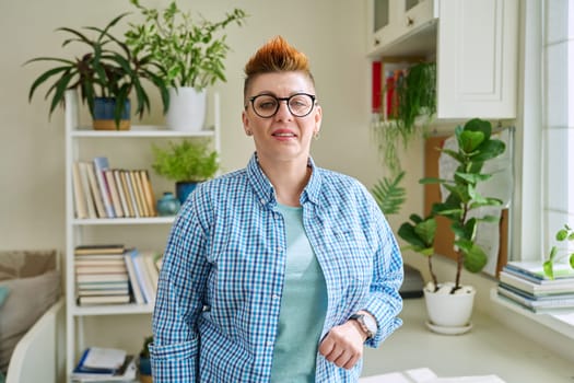 Portrait of smiling middle-aged woman in glasses with red haircut looking at camera in home interior. Mature people, lifestyle, health, life concept