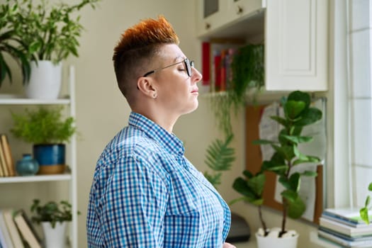 Profile view of middle-aged relaxed woman with closed eyes, at home near window. Calm serene female, relaxation meditation, breathing techniques practice. Lifestyle, health 40s people concept