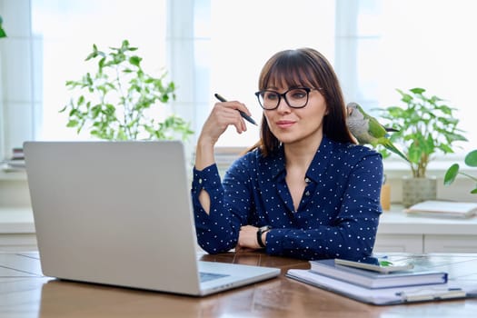 Middle aged woman sitting at workplace with computer in home office, holding pet with green quaker parrot on her shoulder. Work, lifestyle, pets, tropical birds