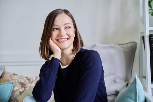 Portrait of young attractive smiling woman in home interior. Happy cheerful relaxed 20s female looking away, sitting on couch. Beauty, youth, happiness, health, lifestyle concept