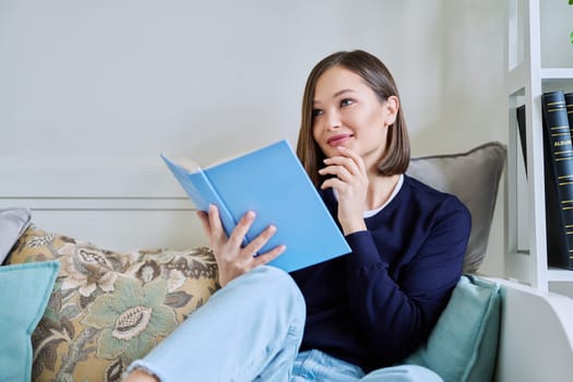 Young woman reading paper book, relaxed sitting on sofa at home. Fiction, hobby, leisure, youth concept
