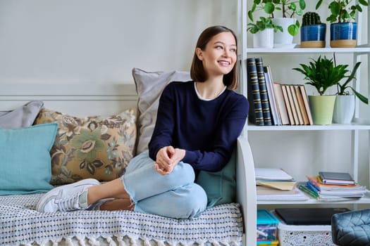 Portrait of young attractive smiling woman in home interior. Happy cheerful relaxed 20s female looking away, sitting on couch. Beauty, youth, happiness, health, lifestyle concept