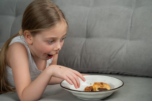 A cute little girl smeared in chocolate eats cookies while lying on the sofa