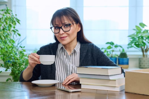Middle aged woman holding cup of coffee, sitting at table with pile of books at home, relaxed smiling looking at camera. Leisure, lifestyle, literary hobby, mature people concept