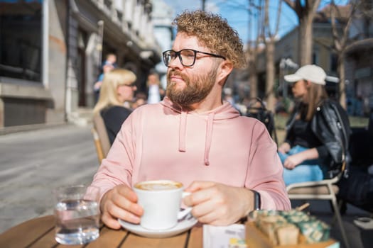 Millennial bearded man having breakfast at table of street cafe on spring day, drinking warm cappuccino. Spring restaurant terrace.