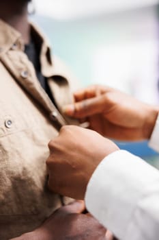 Clothing store assistant hands fastening buttons on customer shirt. African american fashion boutique employee helping client to try on casual apparel before buying with close up on arms