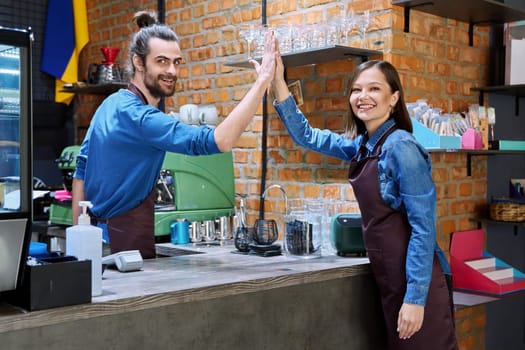 Colleagues young cheerful successful man and woman in aprons give each other five high five gesture at workplace at bar counter in coffee shop cafeteria restaurant. Partnership teamwork small business