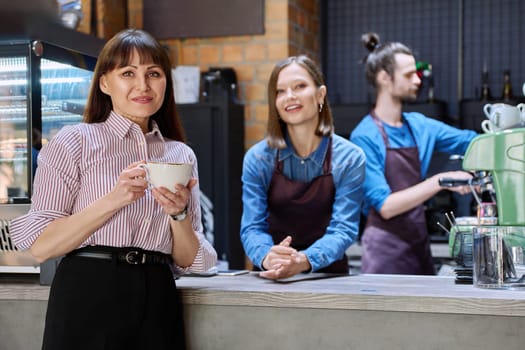 Woman customer of coffee shop near counter with cup of coffee looking at camera, restaurant workers owners at workplace. Colleagues partners in food service work, entrepreneurship, small business