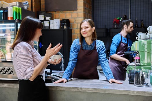Woman customer of coffee shop near counter with cup of coffee talking to restaurant workers owners. Workplace at bar, colleagues, partners in food service, work, entrepreneurship, small business