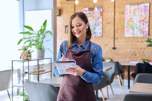 Confident successful young woman service worker owner in apron with working notepad pen in restaurant cafeteria coffee pastry shop interior. Small business staff occupation entrepreneur work