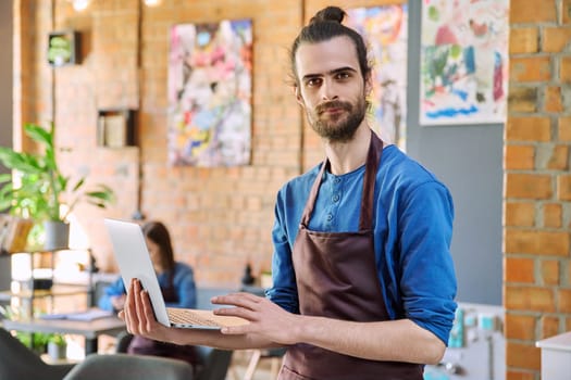 Successful young man service worker owner in apron holding using laptop computer looking at camera in restaurant cafeteria coffee pastry shop interior. Small business staff occupation entrepreneur work