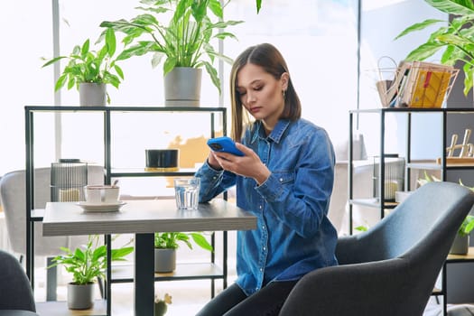 Young attractive woman using smartphone, drinking cup of coffee, glass of water, sitting in cafeteria, cafe. Serious female holding mobile phone, texting messages. Technology lifestyle youth concept