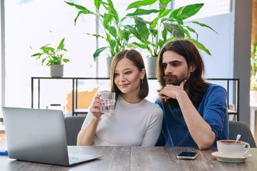 Calm serious young couple looking at laptop together while sitting in cafe, cafeteria. Leisure time for two, lifestyle, togetherness, relationship, communication, work study remotely