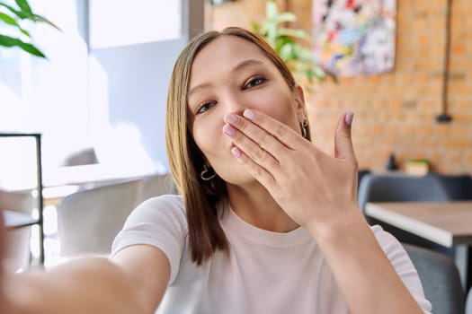 Close up selfie portrait of young smiling attractive woman. Beautiful female female making hand gesture air kiss. Beauty, fashion, youth, lifestyle concept