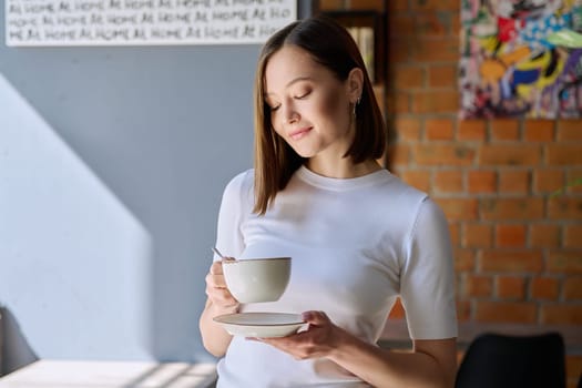Young beautiful happy woman holding cup of coffee tea with saucer looking at cup, gray wall of cafe coffee shop cafeteria copy space for text. Coffee business work services youth lifestyle leisure
