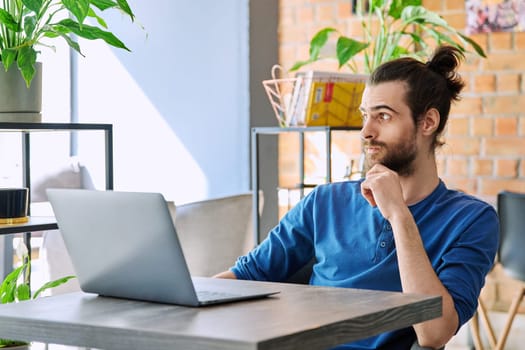 Young serious man working and studying using laptop sitting in coworking cafe. 30s handsome bearded guy looking at computer screen. Technology for leisure work business education communication people