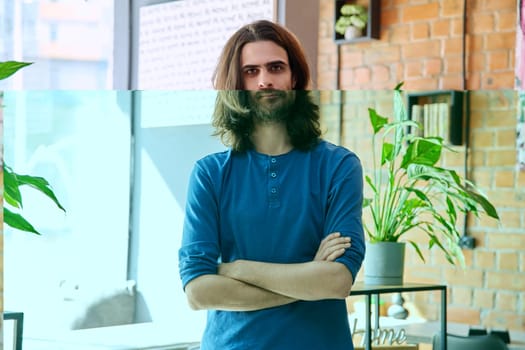 Portrait of young serious confident handsome man with long hair and beard, with crossed arms, looking at camera