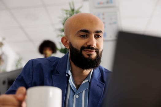 Smiling arab project manager checking customer email on laptop while working in business office. Company start up entrepreneur managing project on computer in coworking space