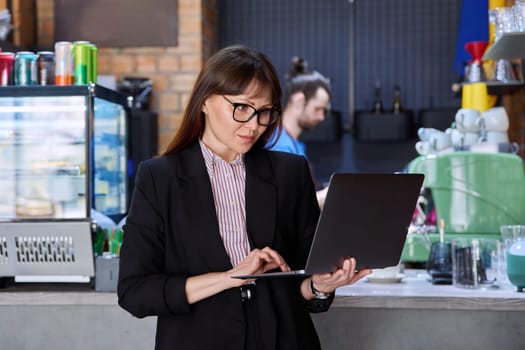 Portrait of confident business woman accountant financier lawyer small business owner. Successful middle-aged female using laptop, looking at computer, background coffee shop cafeteria restaurant