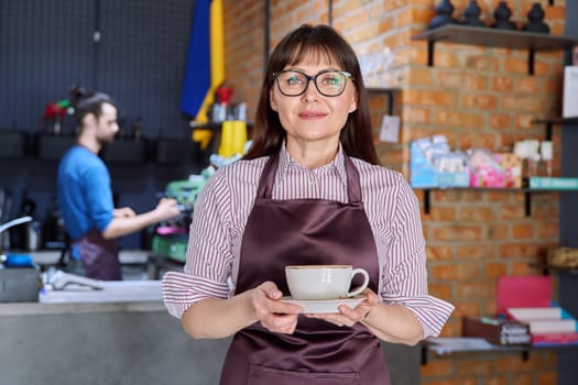 Woman in apron, food service coffee shop worker, small business owner with cup of fresh coffee, looking at camera near bar counter with male barista. Staff, occupation, entrepreneur, work concept