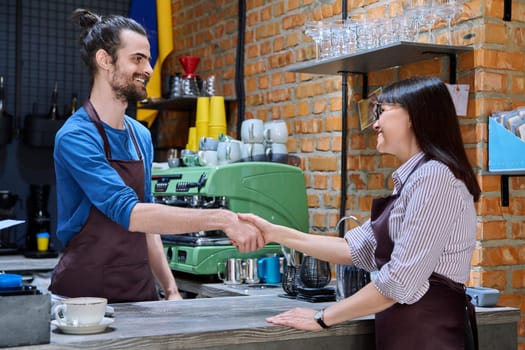 Young man in apron shaking hands with woman owner of coffee shop cafeteria restaurant, colleagues partners working together. Cooperation, small business, partnership, teamwork team, staff, success
