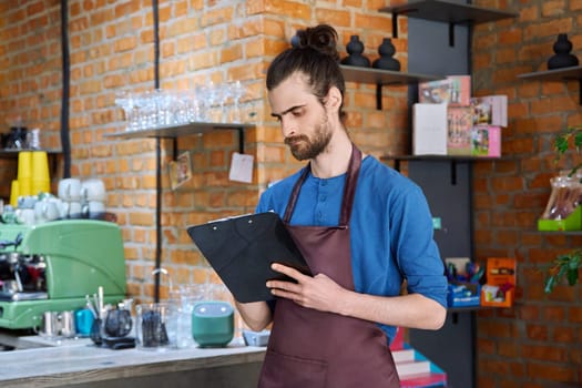 Young serious man in apron, food service worker, small business owner entrepreneur with work papers near counter of coffee shop cafe cafeteria. Staff occupation successful small business work concept