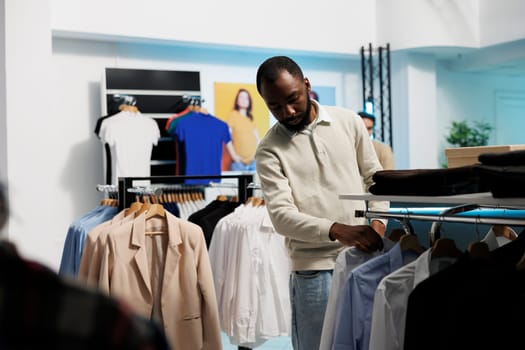 African american man choosing formal outfit and browsing shirts hanging on rack. Young stylish buyer searching for trendy garment while shopping in retail center department store