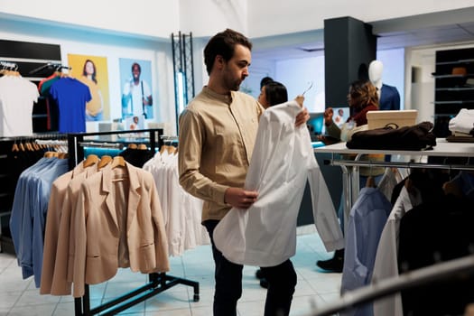 Man holding shirt on hanger, examining style and purchasing casual wear in fashion retail store. African american customer checking formal apparel while choosing outfit in boutique