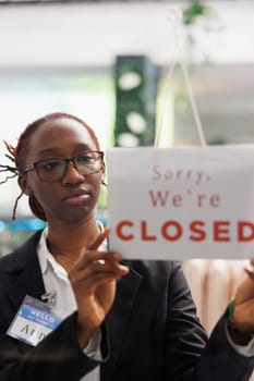 Woman holding closed sign on fashion clothes store front door, informing customers about business hours end. Apparel shop african american assistant hanging sign on widow