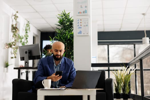 Business arab professional texting on smartphone while working on laptop in start up office. Entrepreneur searching financial information on mobile phone in green open space